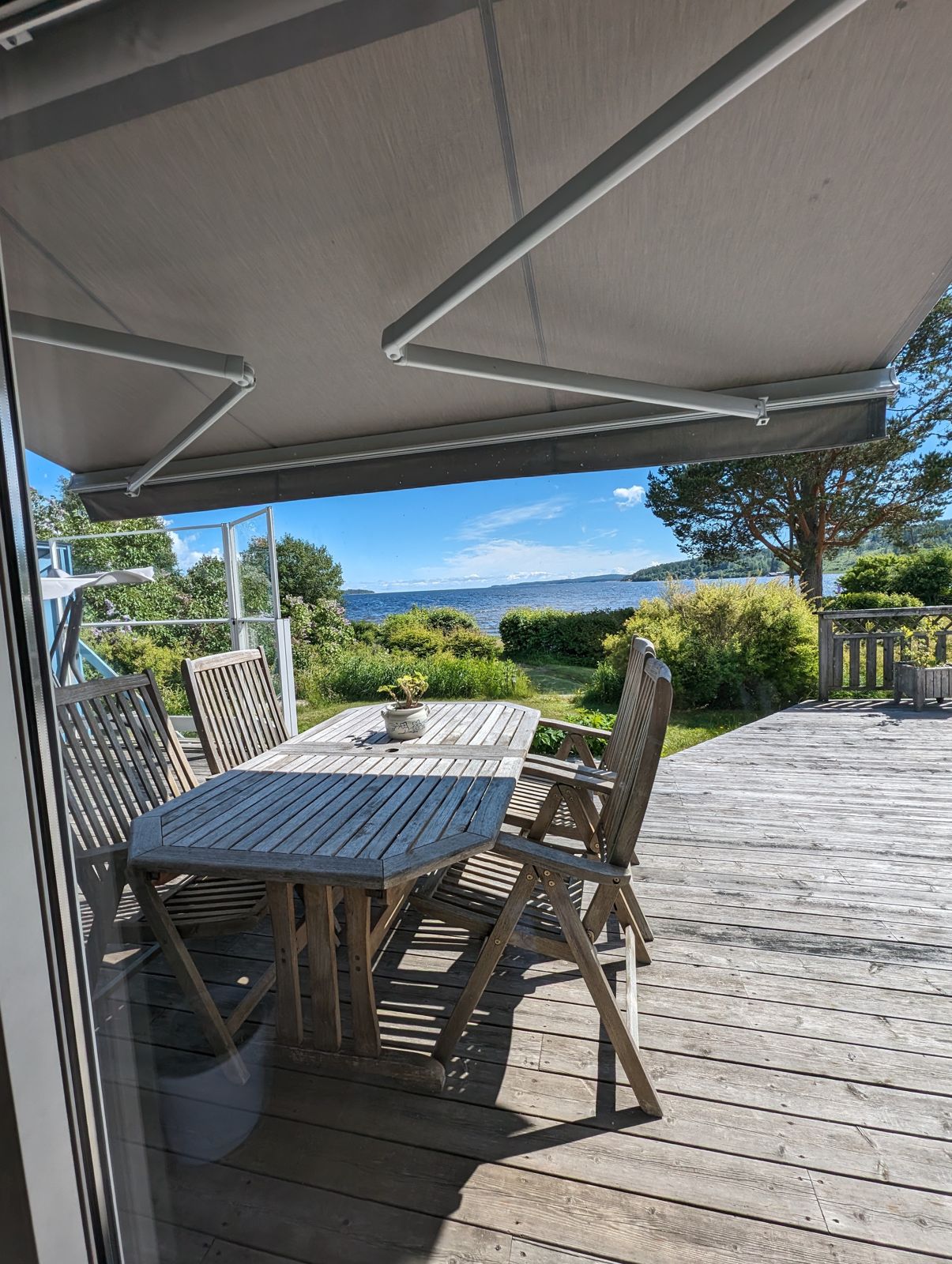 Ljus terrassmarkis över en trätrall med matbord och utsikt mot vattnet