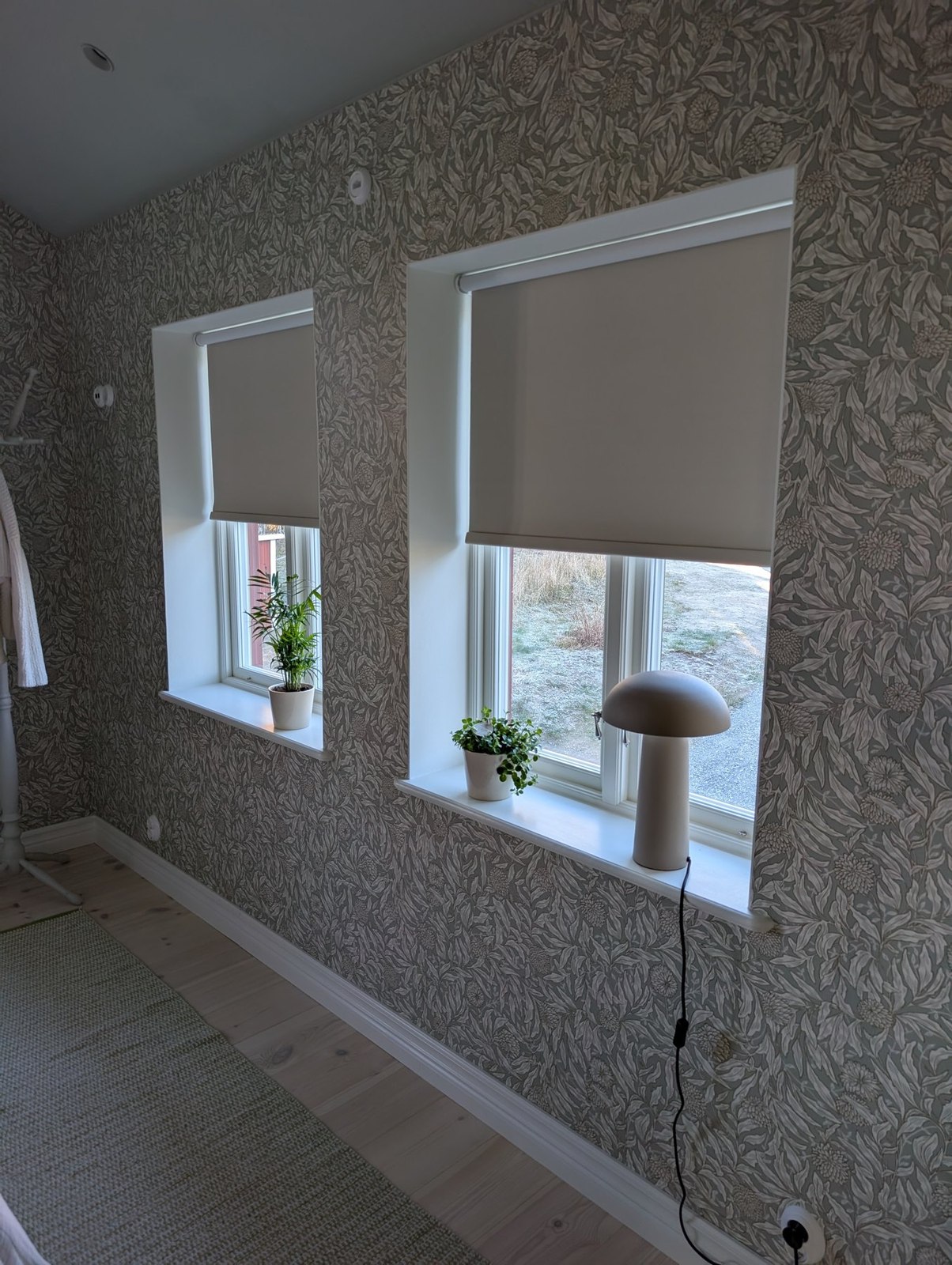 Två fönster med ljusa mörkläggande rullgardiner och blomkrukor i ett sovrum med mönstrad vägg