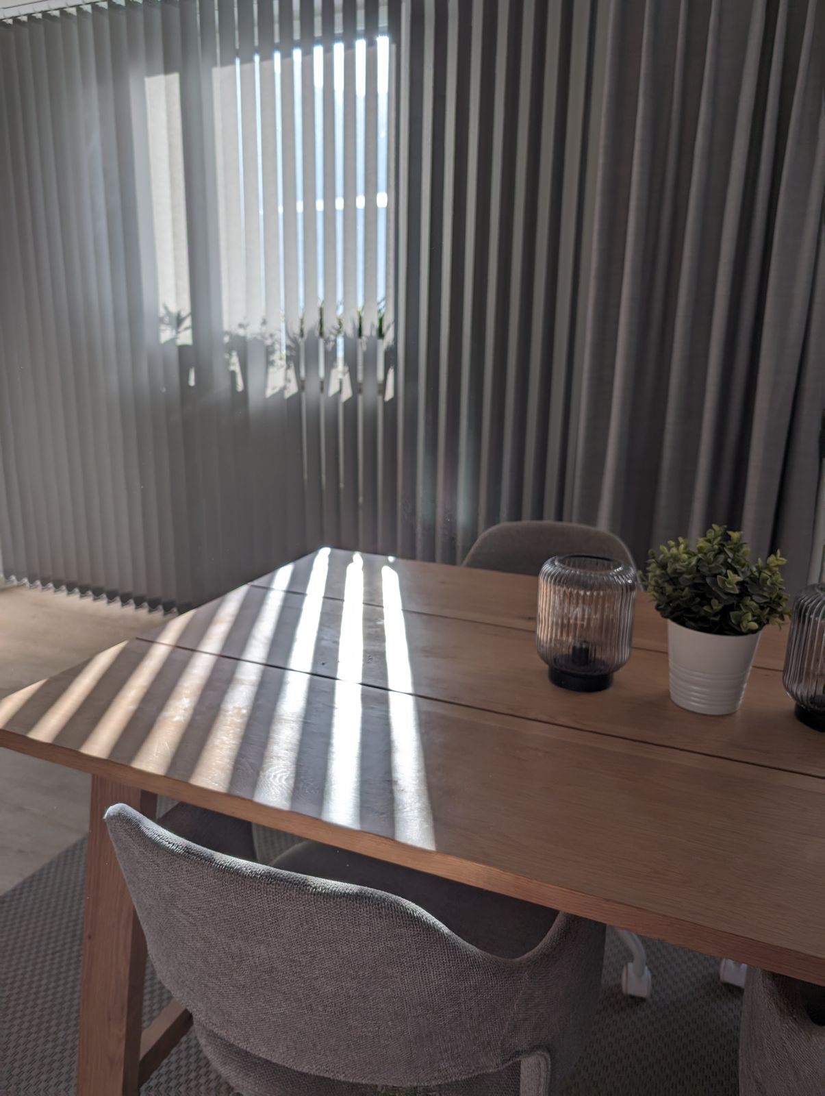Grå lamellgardiner vid matplats där solljus faller i ränder över bordet